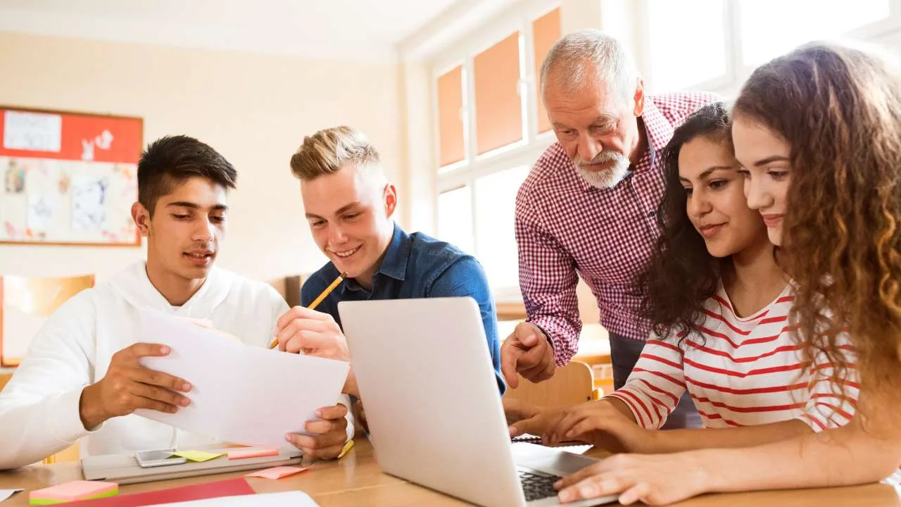 Estudiantes colaborando en un proyecto con un profesor en un aula con una computadora portátil.