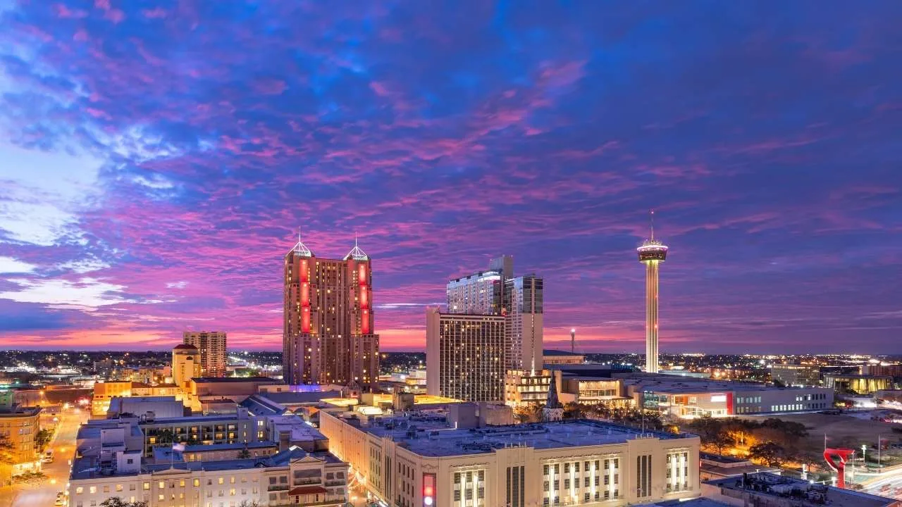 Panorámica de San Antonio con edificios y una torre iluminada durante el atardecer.