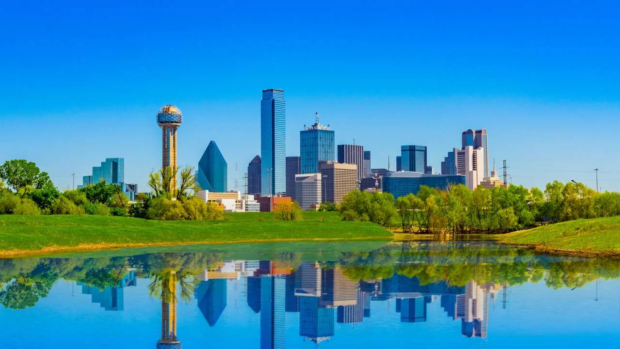 Vista del horizonte de Dallas con edificios y un lago reflejando la ciudad en el agua.