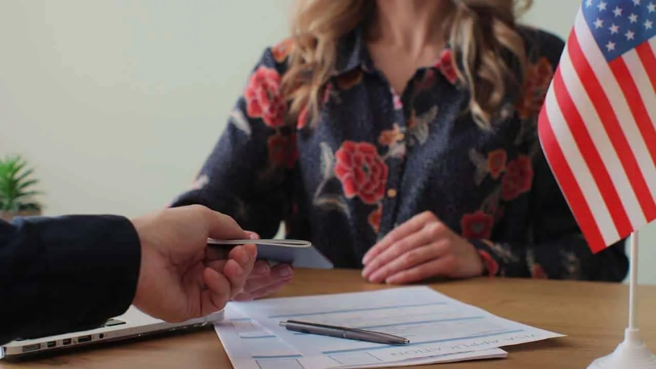 Persona entregando una tarjeta a otra en una oficina de migración con una bandera de EE. UU. en la mesa.