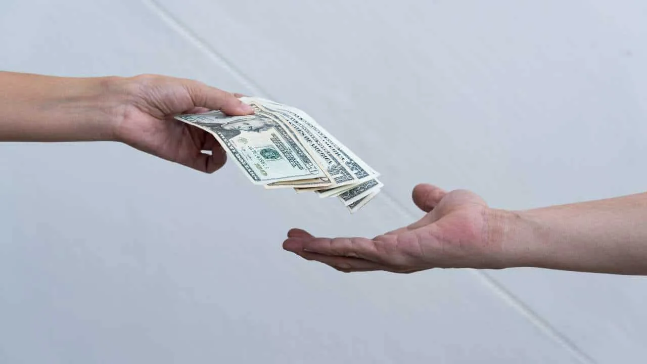 Una mano entrega billetes de dólar a otra mano en un fondo neutro.