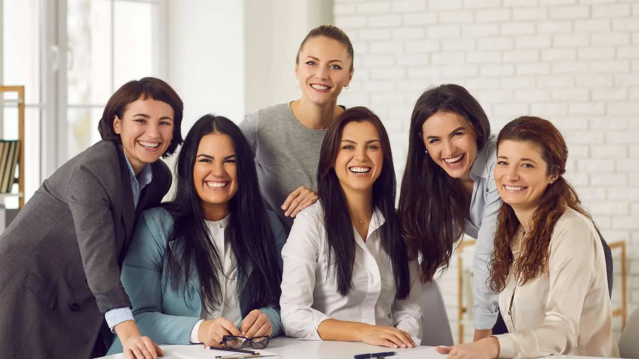 Seis mujeres sonrientes posando juntas en un entorno de oficina con una mesa.