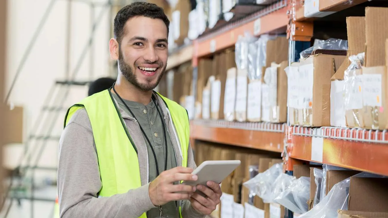 Hombre con chaleco de seguridad sonriendo mientras usa una tablet en un almacén.