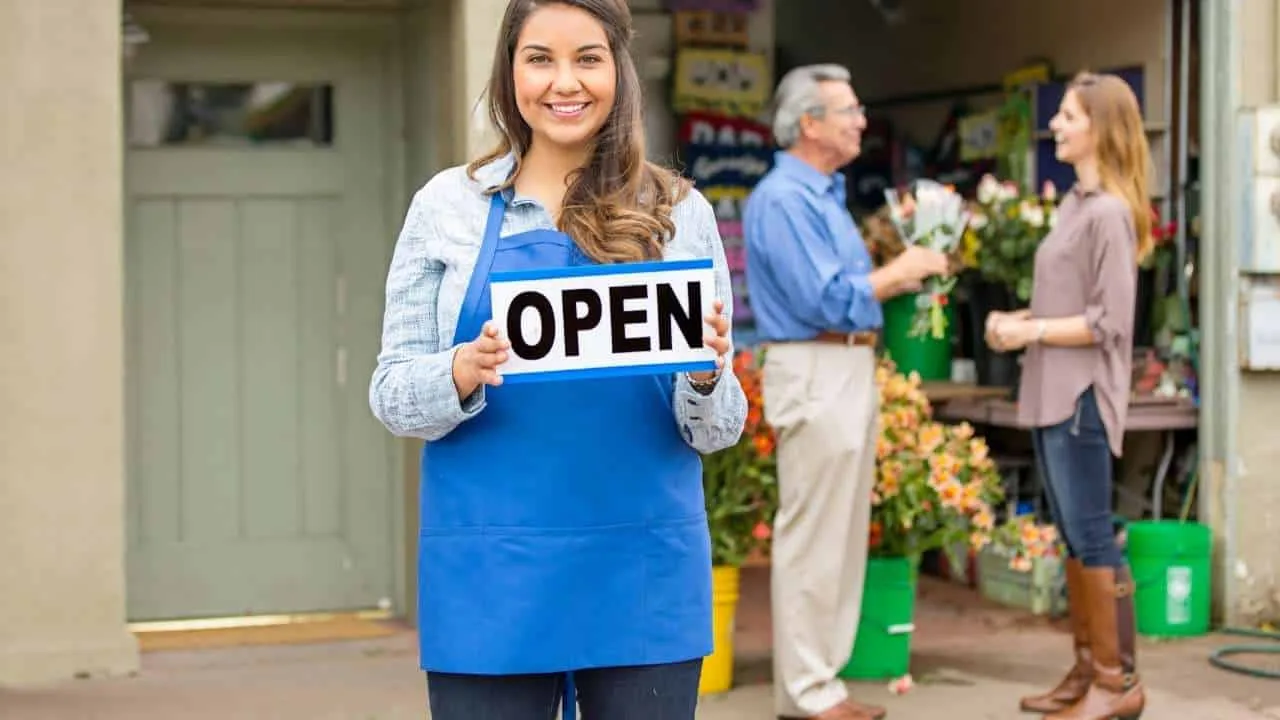 Mujer con delantal azul sosteniendo un cartel que dice 'Abierto' en una tienda de flores.