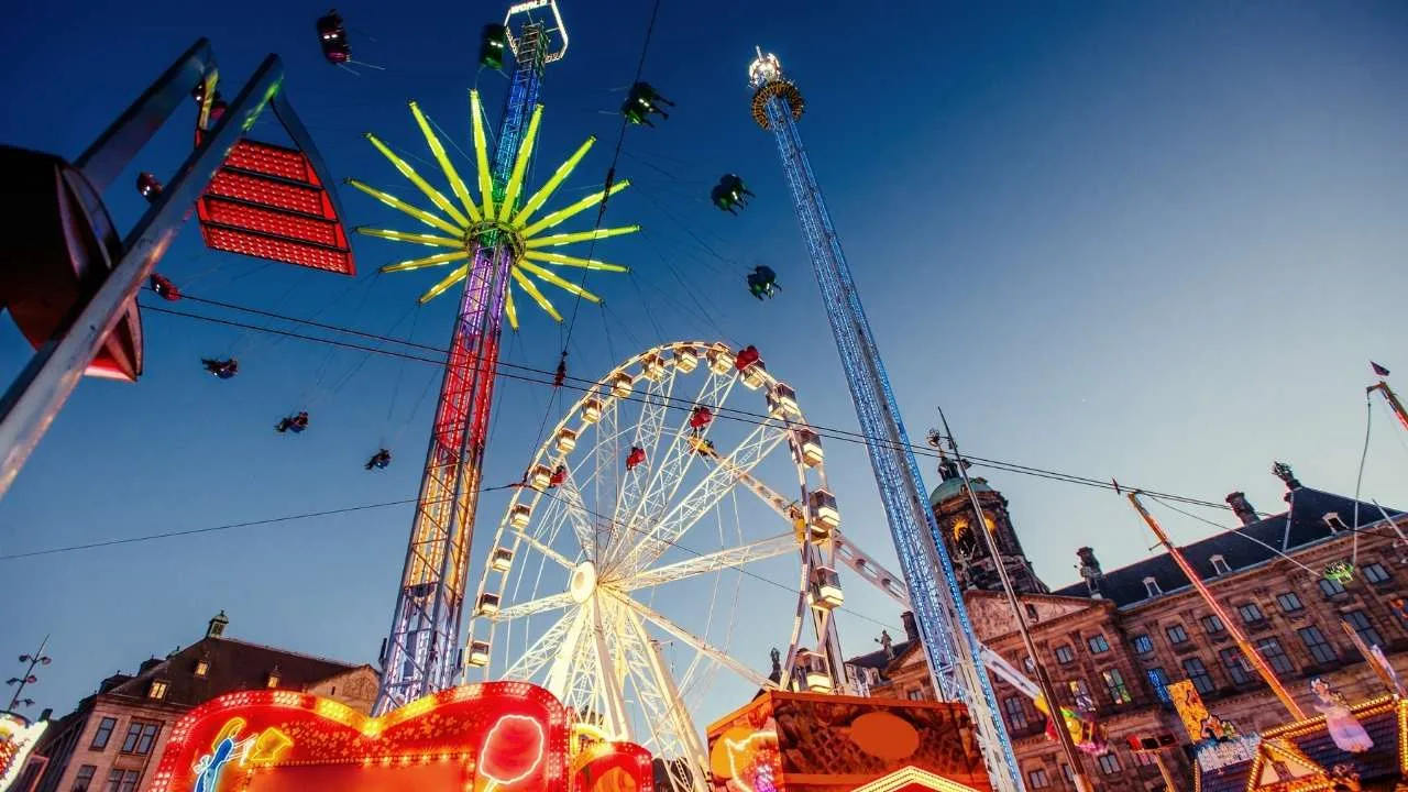 Rueda de la fortuna iluminada y varias atracciones en un parque de diversiones al anochecer.