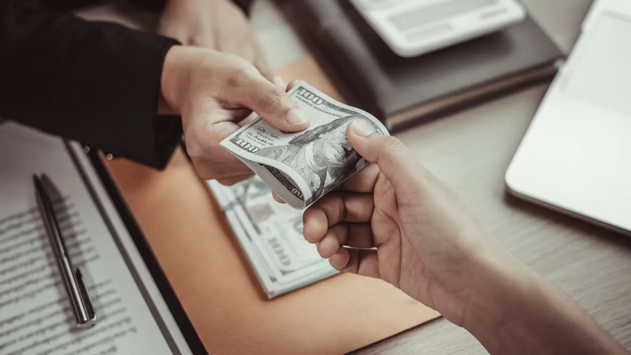 Una mano entrega un billete de cien dólares a otra mano en un entorno de oficina.