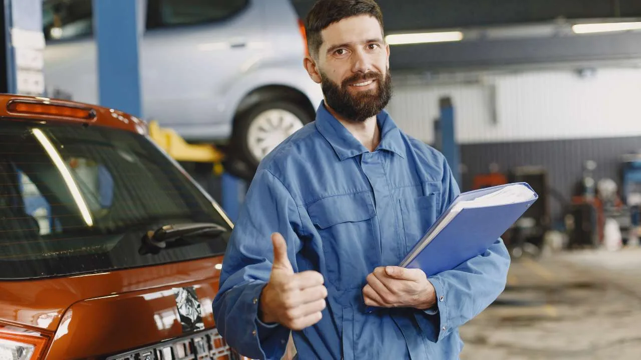 Técnico de vehículos con un cuaderno en mano y haciendo un gesto de aprobación en un taller mecánico.
