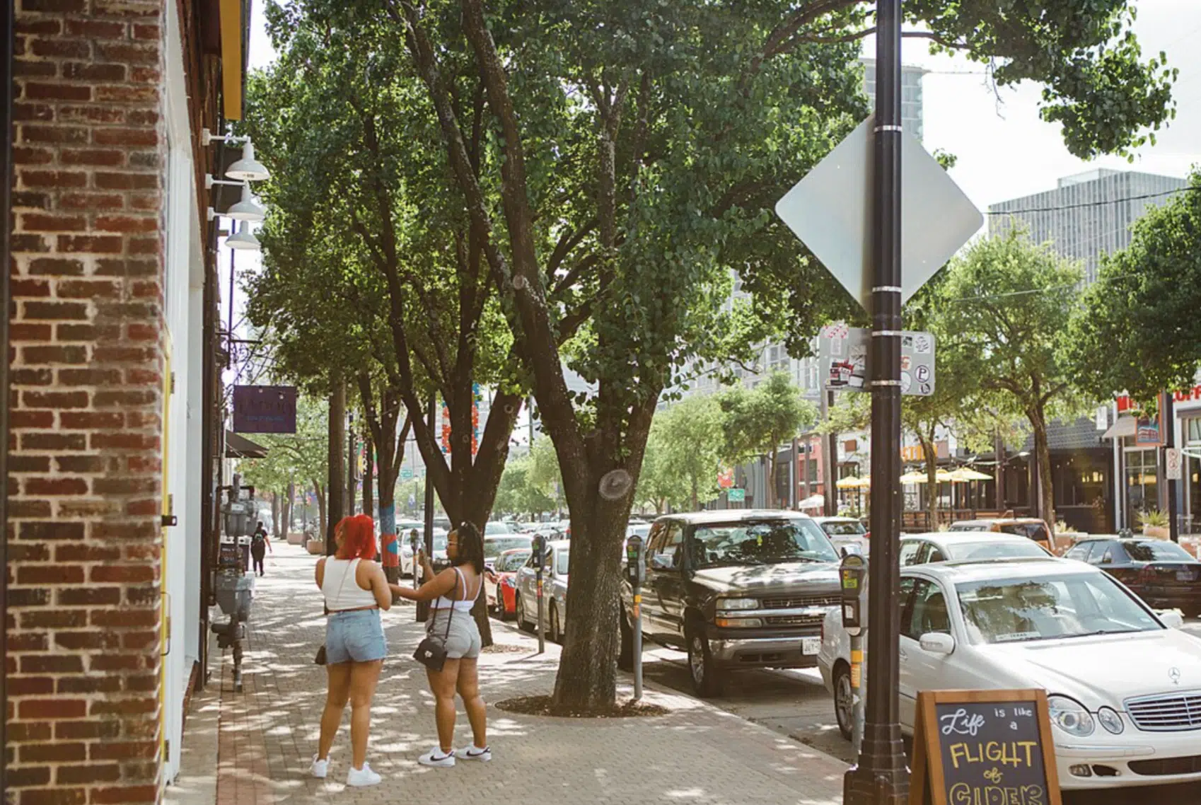 Dos mujeres de pie en una acera arbolada conversando mientras los coches pasan por la calle.