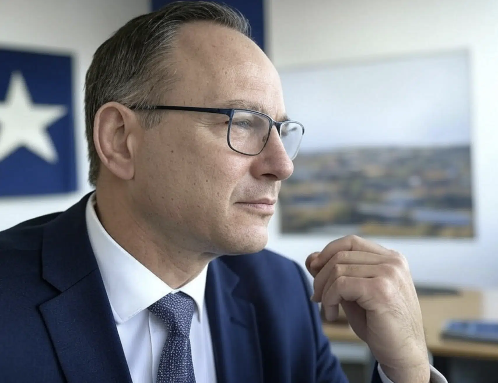 Hombre con gafas y traje oscuro, mirando hacia un lado en un entorno de oficina.