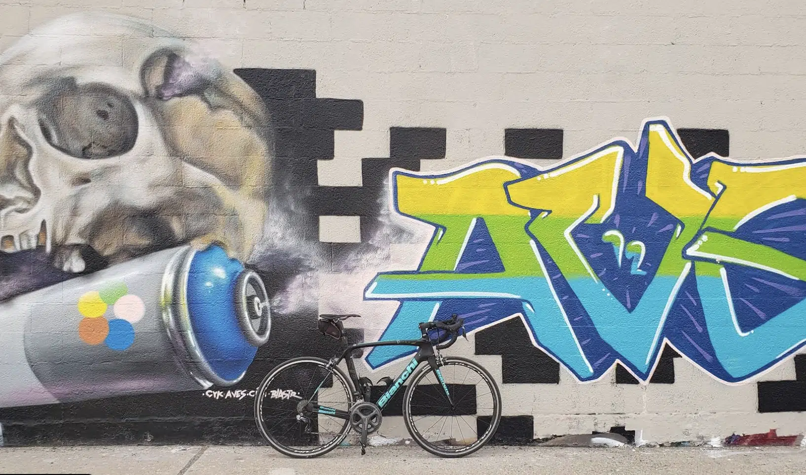 Bicicleta de carretera negra y verde apoyada contra un mural de graffiti con un cráneo y colores vibrantes.