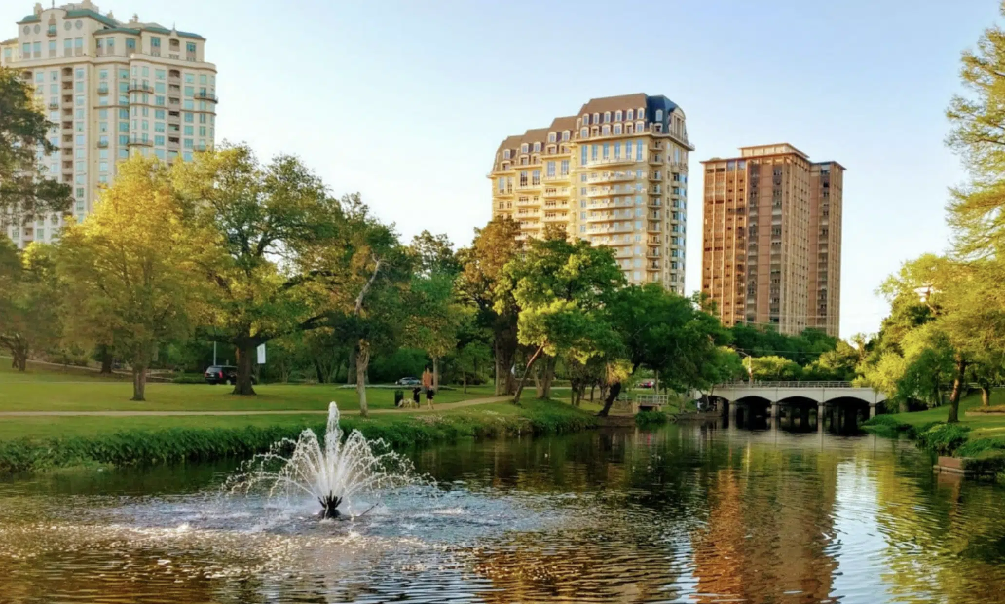 Fuente en un estanque rodeada de árboles y edificios altos en el fondo.